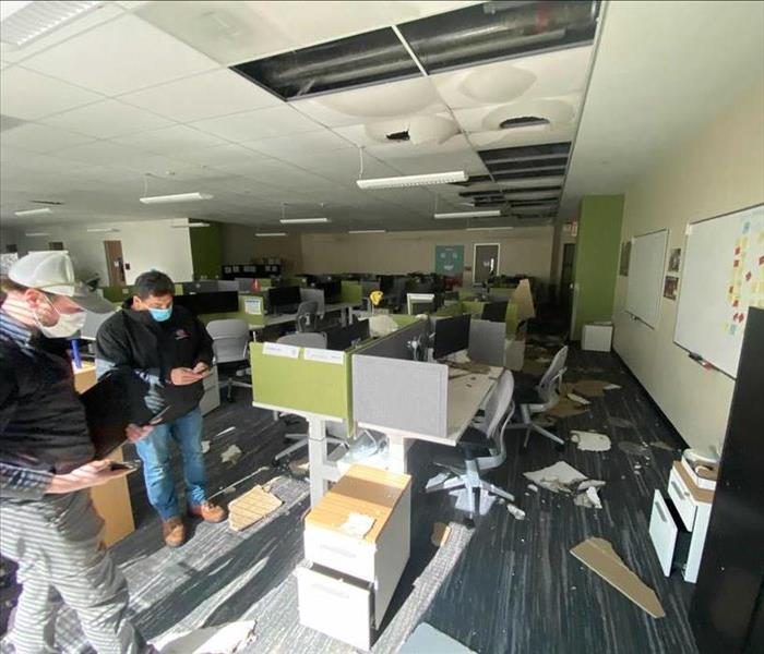 Office area with desk cubicles. 2 men standing in left corner. Ceiling collapsed, debris on the floor and desks