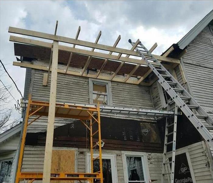 House with ladders and support structure all around it, plywood on one window, side of house falling off on some parts.