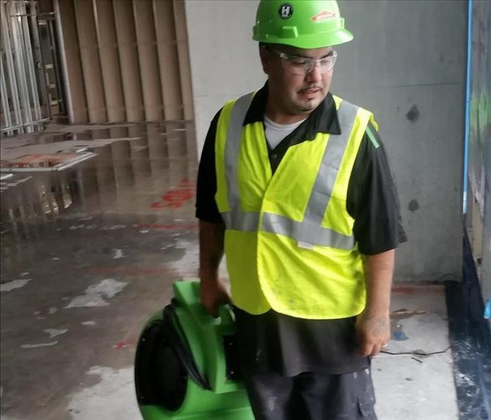 Man standing in bone structure building holding green equipment, wearing yellow safety vest, green helmet, and safety goggles